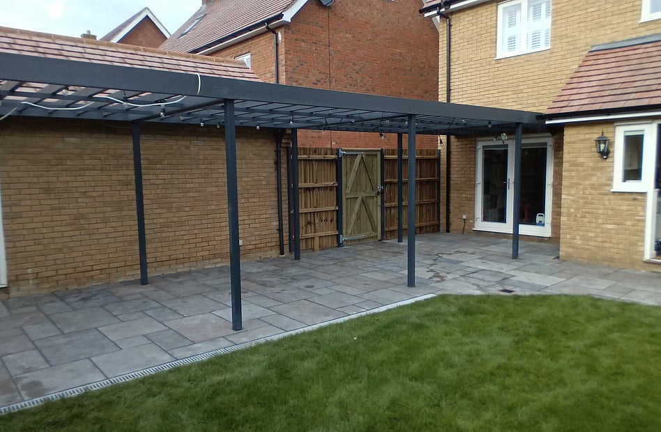 Image shows a completed landscaping project from 2021. It has a sandstone patio with aco drains, new closeboard fencing and gate fitted using Durapost components. Above the patio is a pergola, utilising Durapost gate posts and finished with soft wood timber rafters and painted with Anthracite coloured paint.