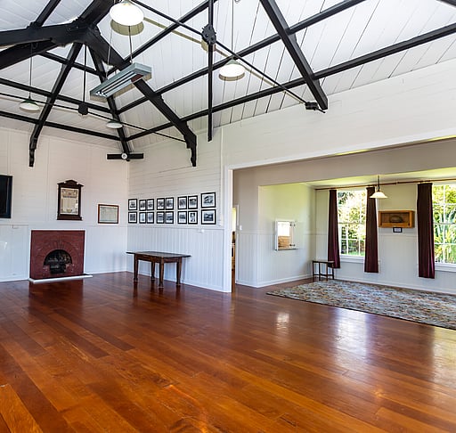 An interior view of the Kūaotunu Hall showing the main part of the hall and the annex on the right hand side. The floor of the hall is swamp kauri and the walls are white, there are two tall sash windows with red velvet curtains.