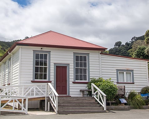 The Kūaotunu Hall as seen from the front - a white painted wooden building with a red roof and red door, with steps leading up to it and a ramp to the left