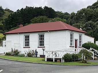 The Kūaotunu Hall in 2023 - a white wooden building with a red roof, with steps up to the front door and a ramp to the left side