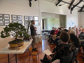 The Kūaotunu Garden Group listening to a speaker in the Kūaotunu Hall who is standing alongside a table on which are examples of bonsai trees