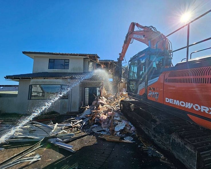 Excavator demolishing a house while a water spray keeps the dust down