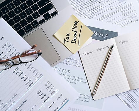 Laptop with Post-it note stating Tax Deadline and lots of paperwork lying around it, showing numbers and reports. Pencil and notepad.