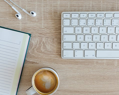 Wooden table with a Notebook, Coffee Mug, white keyboard and Earbuds laying on the table.