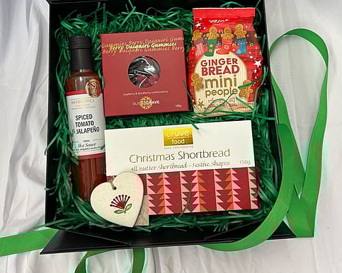Christmas Treat Gift Box with Herb & Spice Hot Sauce, Sugarcrave Gummies, Bisko Gingerbread Mini People, Crave Shortbread, and Michelle Bow Ceramic Heart Ornament in black gift box with green filler.
