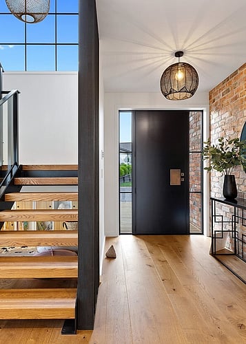 A modern entranceway with black front door, brick wall, wooden stairs and a natural wood floor. 