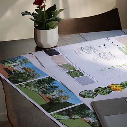 A garden concept plan is laid out on a wooden table. There's a small potted plant in the background.