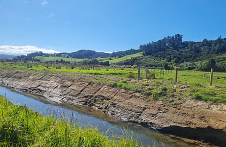 Rural landscape with grassy paddocks, fence line and narrow stream running through farmland under clear blue sky
