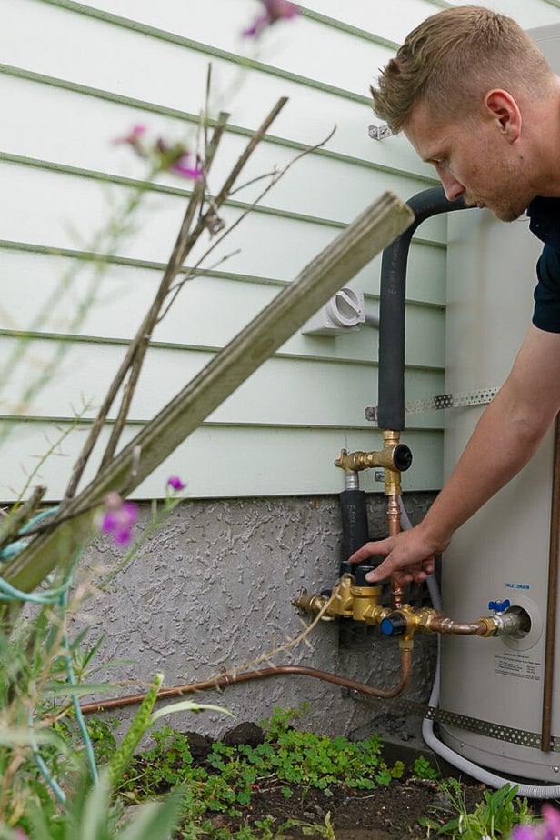 man leaning over tightening valve on exterior hot water cylinder, a plus plumbing and gas logo visible on his shirt, brass pipework visible entering side of house