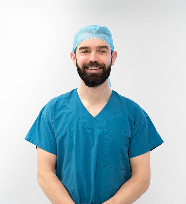 Hamish McLaren n an operating room, wearing surgical scrubs and a cap, operating a robotic surgical device while another medical professional works in the background.