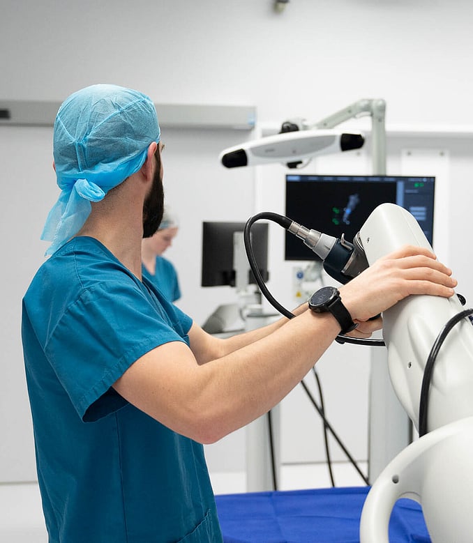 Hamish McLaren n an operating room, wearing surgical scrubs and a cap, operating a robotic surgical device while another medical professional works in the background.