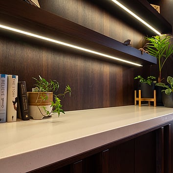 White desk with dark timber wall and built in shelving with lighting. 