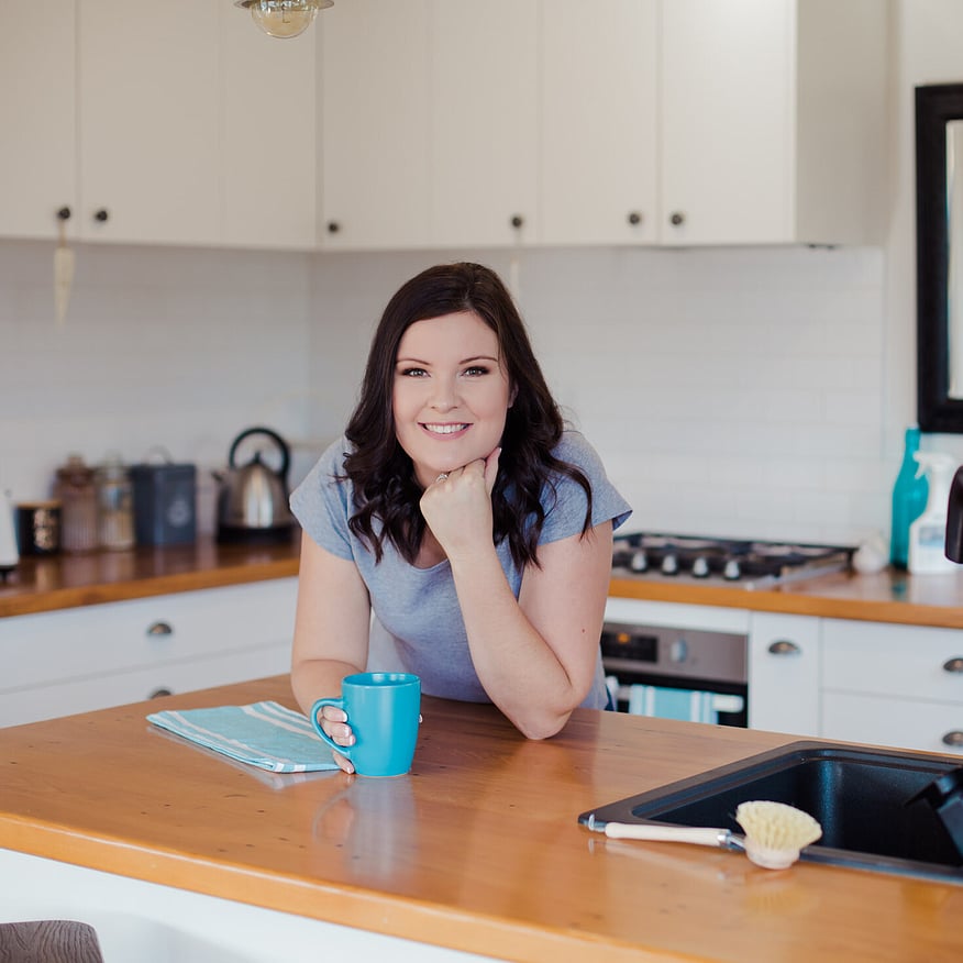 Smiling woman with dark hair leans on a wooden kitchen bench, holding a blue mug, with a sink and cooking essentials in the background