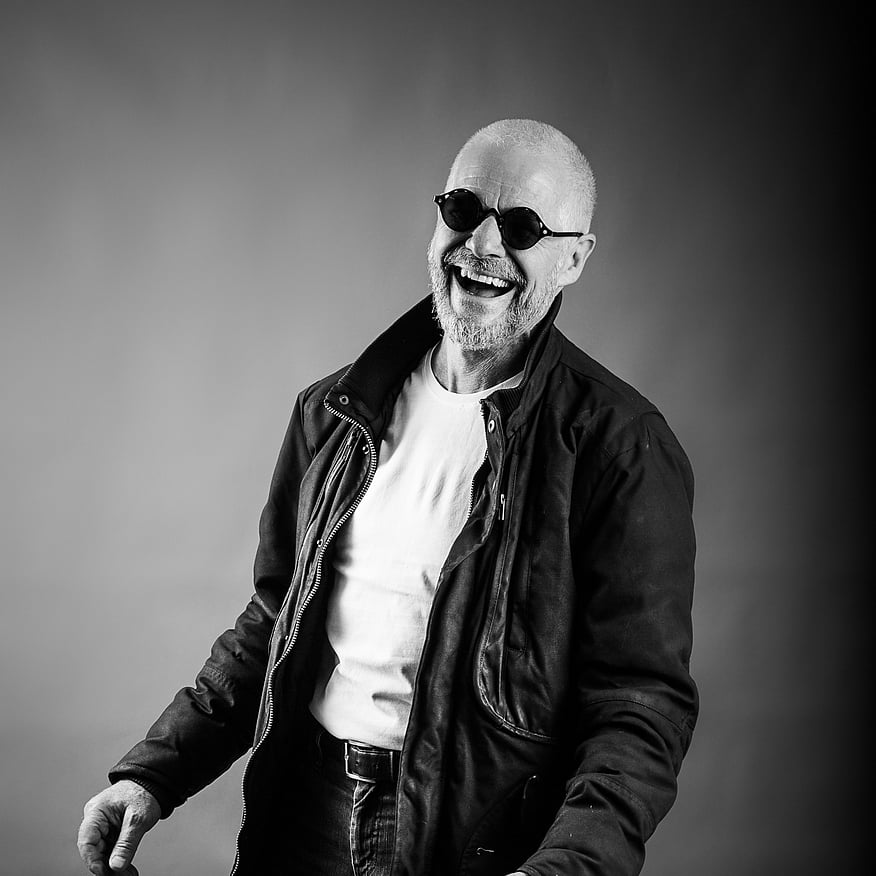A man in a black leather jacket and sunglasses laughing and posing in front of a neutral backdrop, captured in black and white