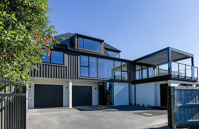 The image shows a newly completed builder's renovation in Papamoa. The home is two two-story with white textured coating and a twin garage on the lower level, and on the upper level, a new deck with pergola, profiled black metal cladding, and large windows. The image is taken from the driveway.