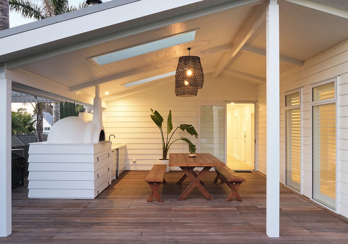 An outdoor kitchen at night with warm lighting illuminating the space. The outdoor kitchen sits on hardwood decking and shows a wooden table, pizza oven, and kitchen units. The area is covered by a beautiful raked ceiling.