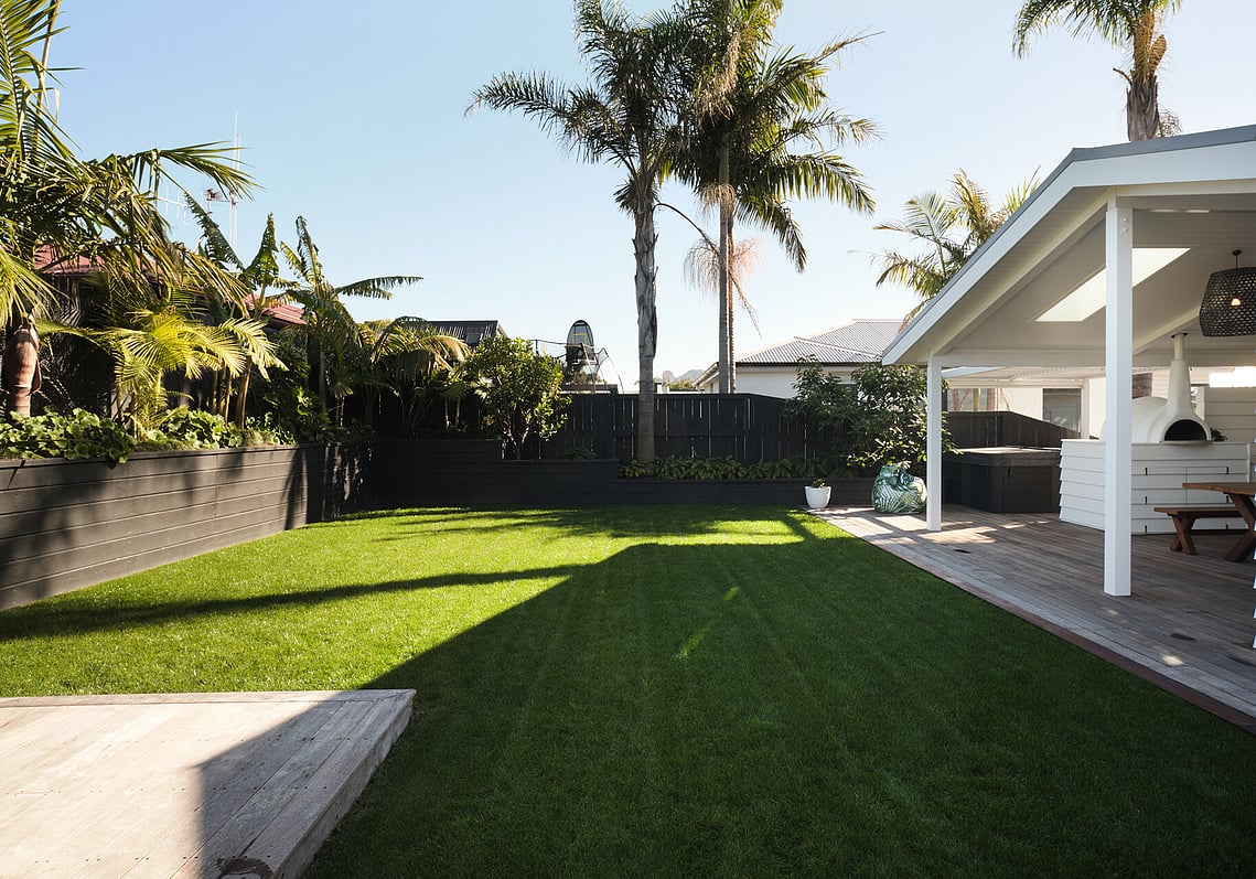 An outdoor garden setting showing the outdoor kitchen areas to the right and the corner of the deck on the left. Immaculate green lawn, raised garden, fence, and palm trees. 