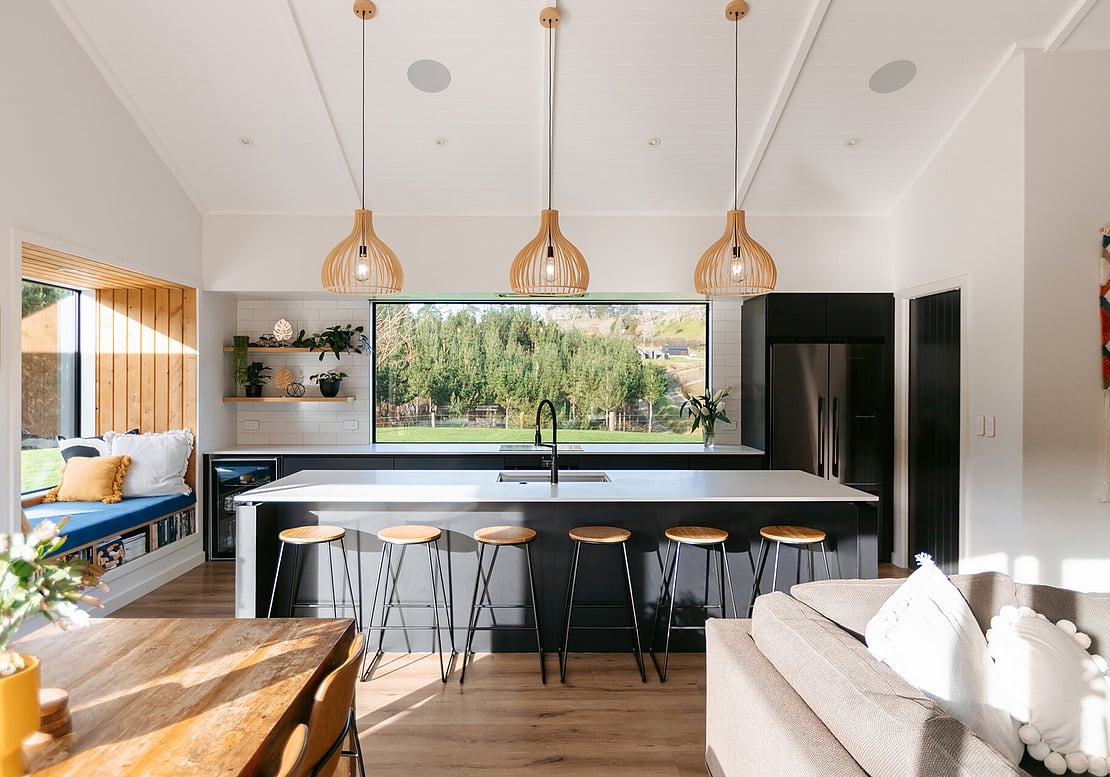The image shows the new kitchen and open-plan dining lounge area in a modern new home in Tauranga. There is also a new window seat to the left of the kitchen. Modern pendant lights hang over the kitchen island. 