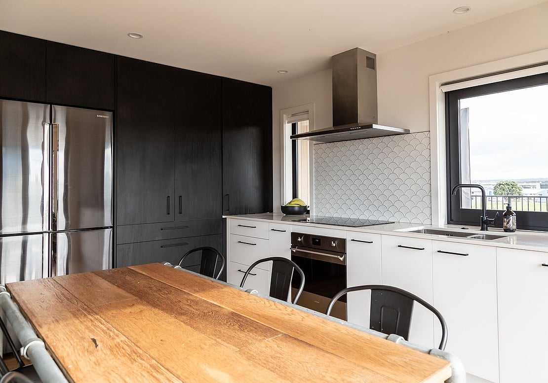 The image shows a newly completed kitchen renovation, including a splashback, white kitchen units, full-height black kitchen units on the far wall, and a fitted chrome fridge. The kitchen window overlooks Simpson Reserve in Papamoa. 