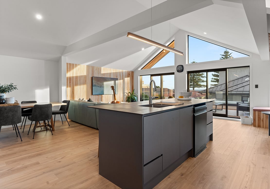 This image shows a newly renovated kitchen, lounge, and dining area with new flooring, internal walls, paint, lights, and a kitchen island with a sink. The style is clean and modern, and the workmanship is completed to a very high standard by local Mount Maunganui Builders called Straight Up Builders Tauranga. 