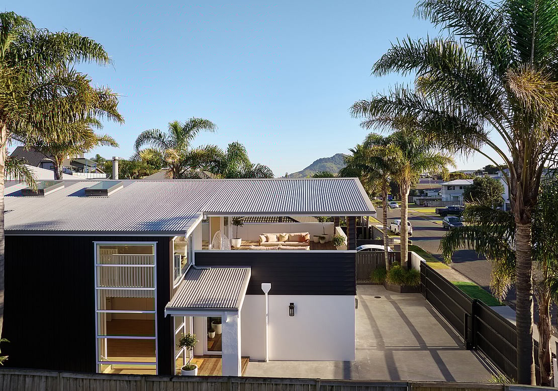 A full home renovation in Mount Maunganui, showing a modern home with a covered deck, surrounded by palm trees, with Mount Maunganui (Mauao) in the distance. 