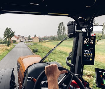View from inside the Valtra S Series cab, showing a farmer&rsquo;s hands on the steering wheel, driving down a rural road with the SmartTouch interface and expansive front visibility.