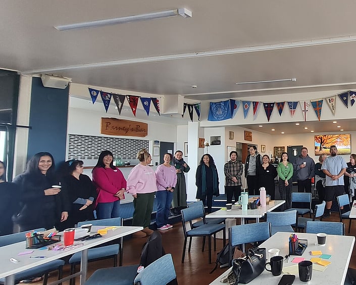 Group of people standing in a circle in a community space, with tables, chairs and notes set up for a collaborative workshop.