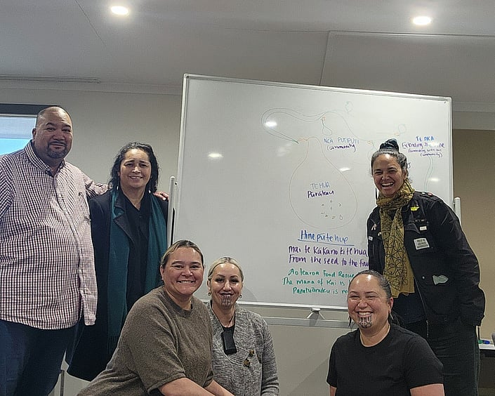 Group of people smiling in front of a whiteboard with a Māori strategy diagram, showing a seed to fruit concept and community connections.