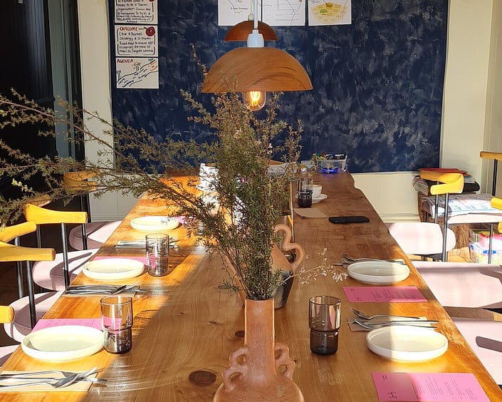 Long wooden table set for a shared meal, with plates, cutlery, glasses and a vase of greenery under warm lighting.