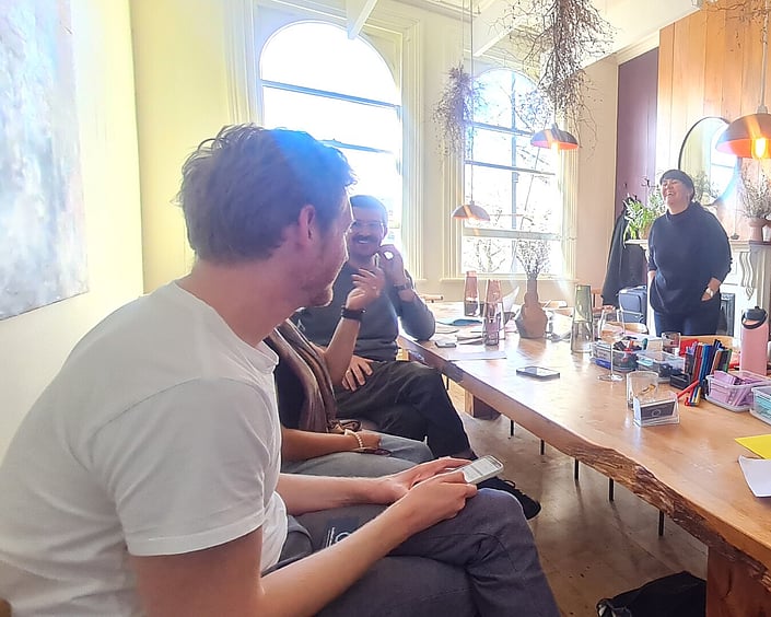 People chatting and laughing around a table in a bright room, with notebooks, pens and drinks spread out during a workshop session.