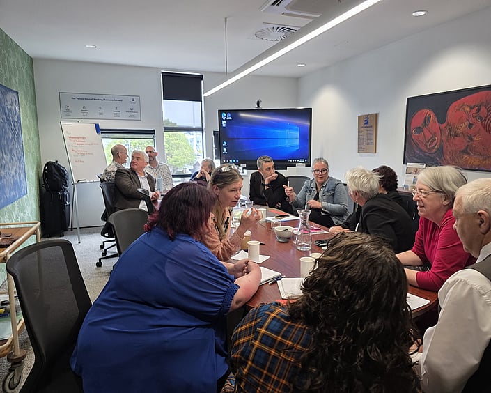 Group of people gathered around a meeting table, discussing ideas during a workshop with notes, drinks and a screen in the background.