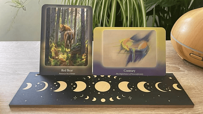 Two cards from the council of horses Oracle card deck on a moon rectangular shaped wooden Oracle card holder with plants and a diffuser in the background