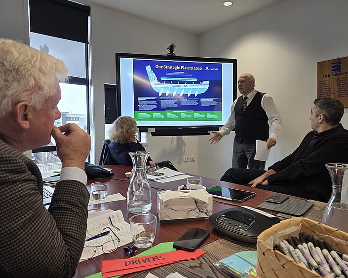 Person presenting a strategic plan on a screen to a small group in a meeting room, with notes, devices and drinks on the table.