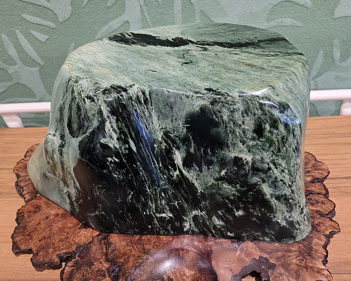 Polished green stone with natural patterns and textures, displayed on a wooden base in an indoor setting.