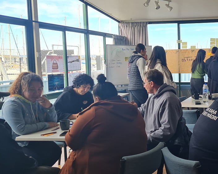 People working together at tables during a workshop, writing notes while others stand by planning boards in a bright room.