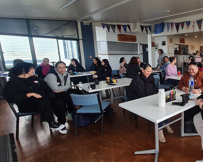 Group seated at tables in a community room, chatting and participating in a workshop with notes, pens and drinks on tables.