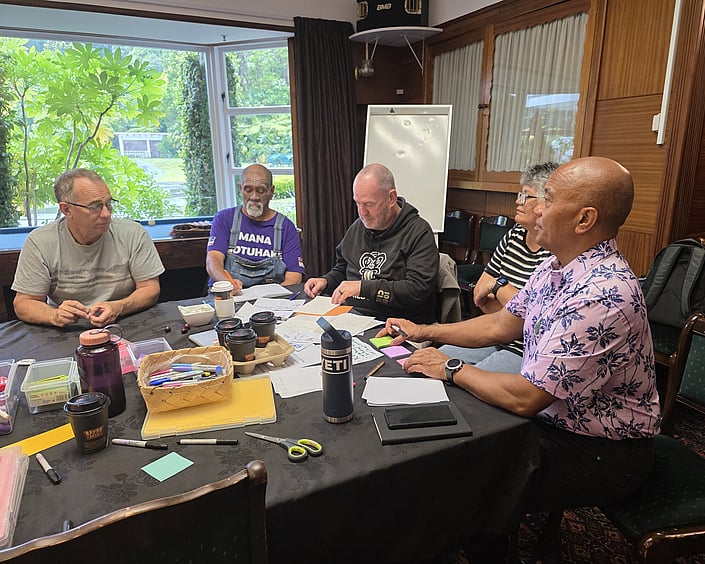 Small group seated around a table discussing strategy with notes, tools, and materials spread out.