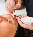 A beauty therapists hands one is holding a dish with eyebrow tint in. The other hand is applying the tint to a lady's eyebrow