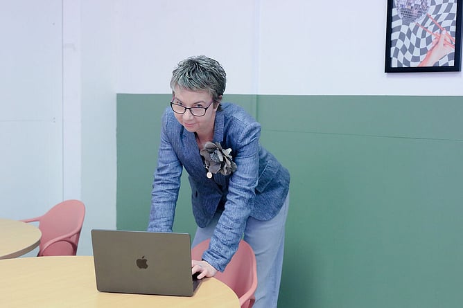 maree gately marketing strategist leaning over her laptop on a table looking into the camera; wearing a jacket with a flower brooch and jeans. Background wall is green and a peach chair.