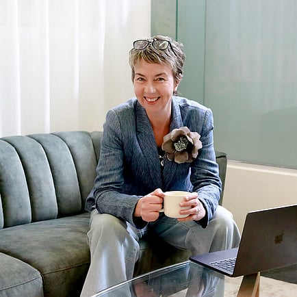 maree gately marketing consultant in whangarei nz sitting on a couch with a coffee in her hand smiling, and wearing jeans and a blazer with a flower on the lapel