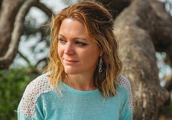 Portrait of a female wedding stationery designer sitting outdoors beside a tree, wearing a soft blue knit top and looking thoughtfully into the distance. Natural, calm lifestyle image representing the balance between motherhood, creativity, and running a flexible creative business.