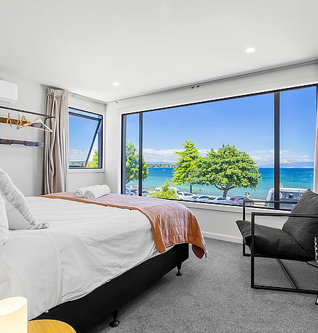 King bed with lake views through floor-to-ceiling windows, neutral linen, and tan throw