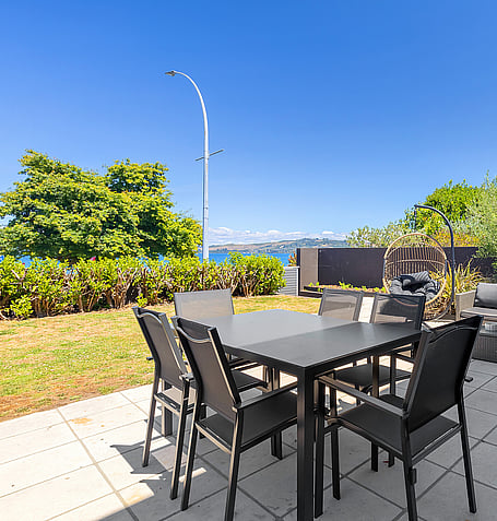 Black outdoor dining table and six chairs on tiled patio with garden and lake glimpse beyond