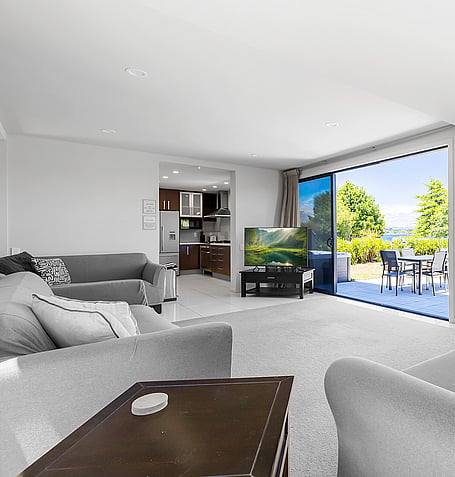 Grey corner sofa and coffee table facing TV, with kitchen and lake-view deck in the background
