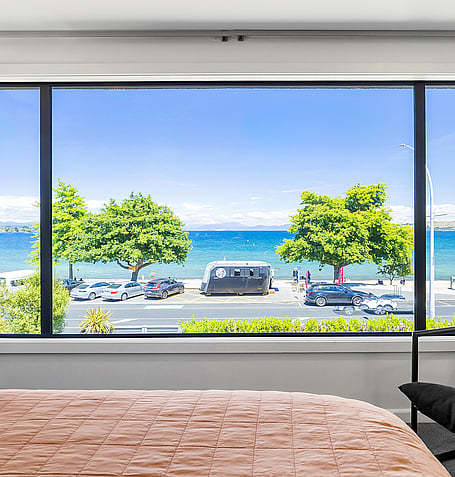 Lake Taupo and waterfront road seen through bedroom window, with tan bed linen in foreground