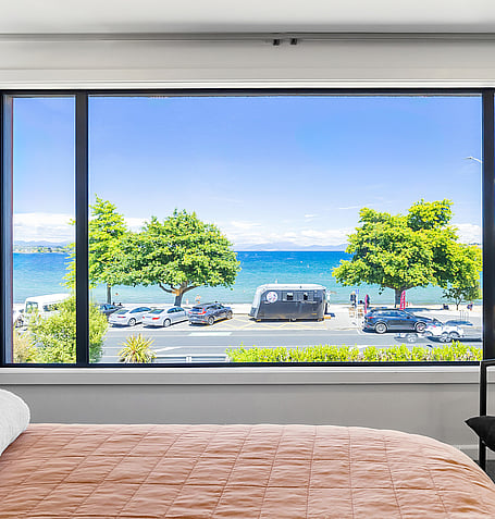 Lake Taupo and waterfront road seen through bedroom window, with tan bed linen in foreground