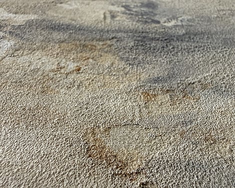 detail showing granular mineral surface with layered warm cream and stone tones, soft grey sweeps, raw sienna markings with exposed pockets of linen underlayer