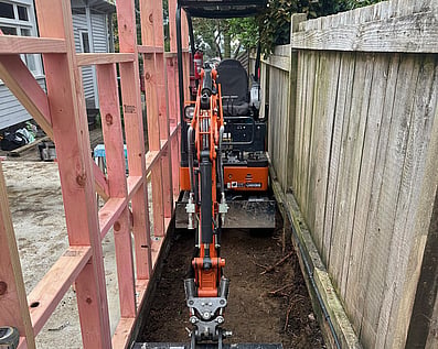 Orange tight-access digger navigating a narrow residential. boundary fence during an excavation project in Auckland.