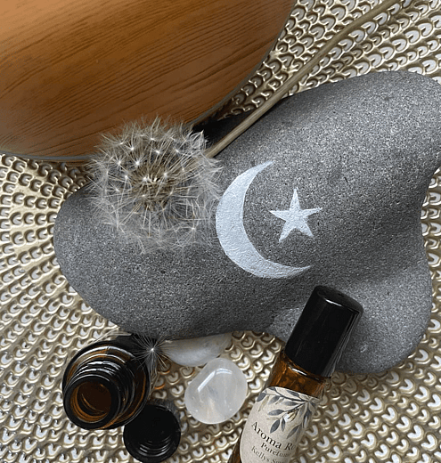 a stone with a moon and star, a dandelion, some essential oil bottles and a crystal on a gold placemat with a diffuser in the background