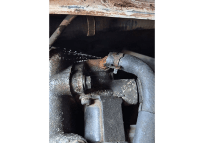 Close-up of a car engine with coolant spraying from a corroded hose fitting. The metal around the joint is rusted, showing a cooling system leak caused by failed anti-freeze protection.
